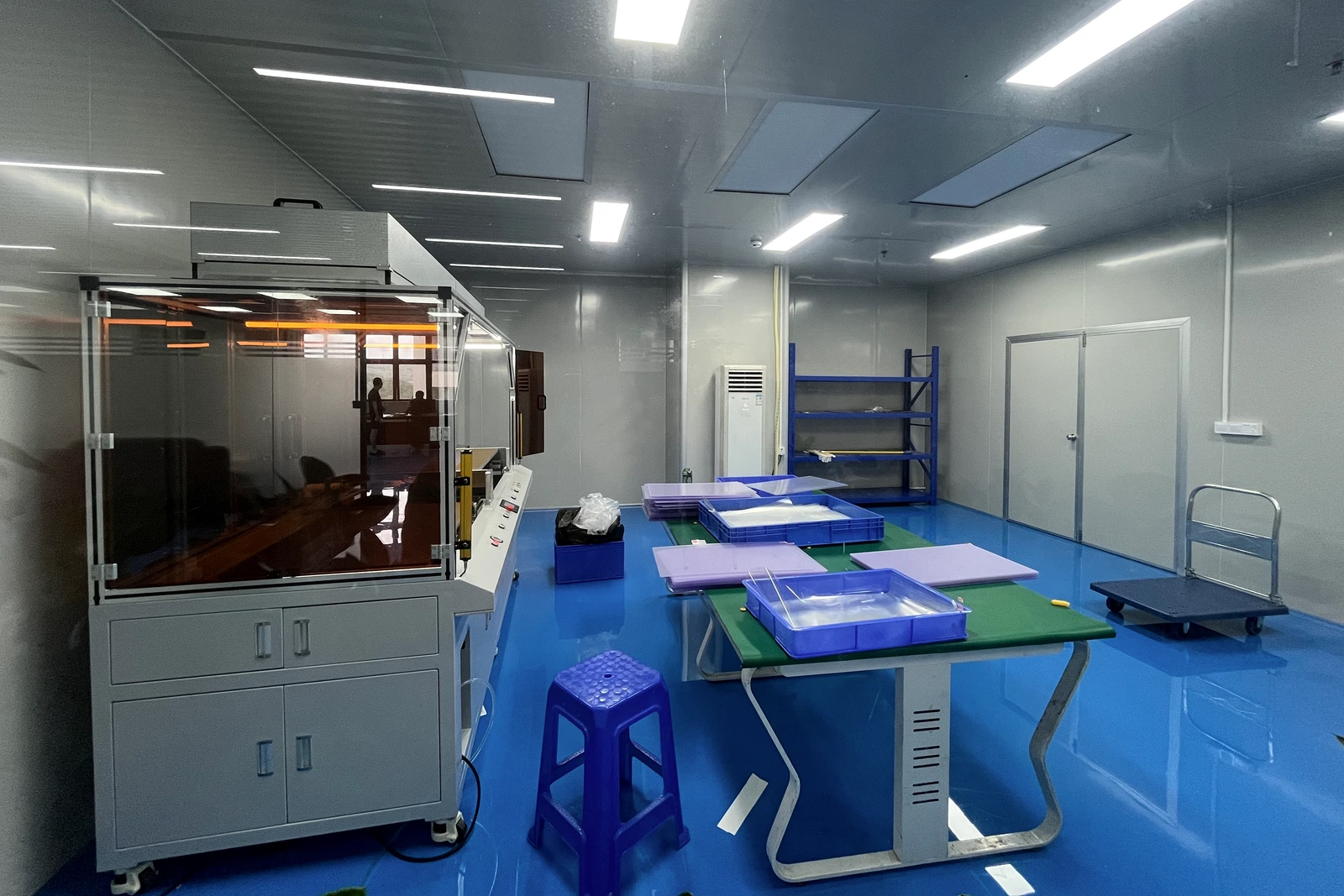 Enclosed equipment stands beside worktables, trays, sheet materials and a trolley inside a production room.