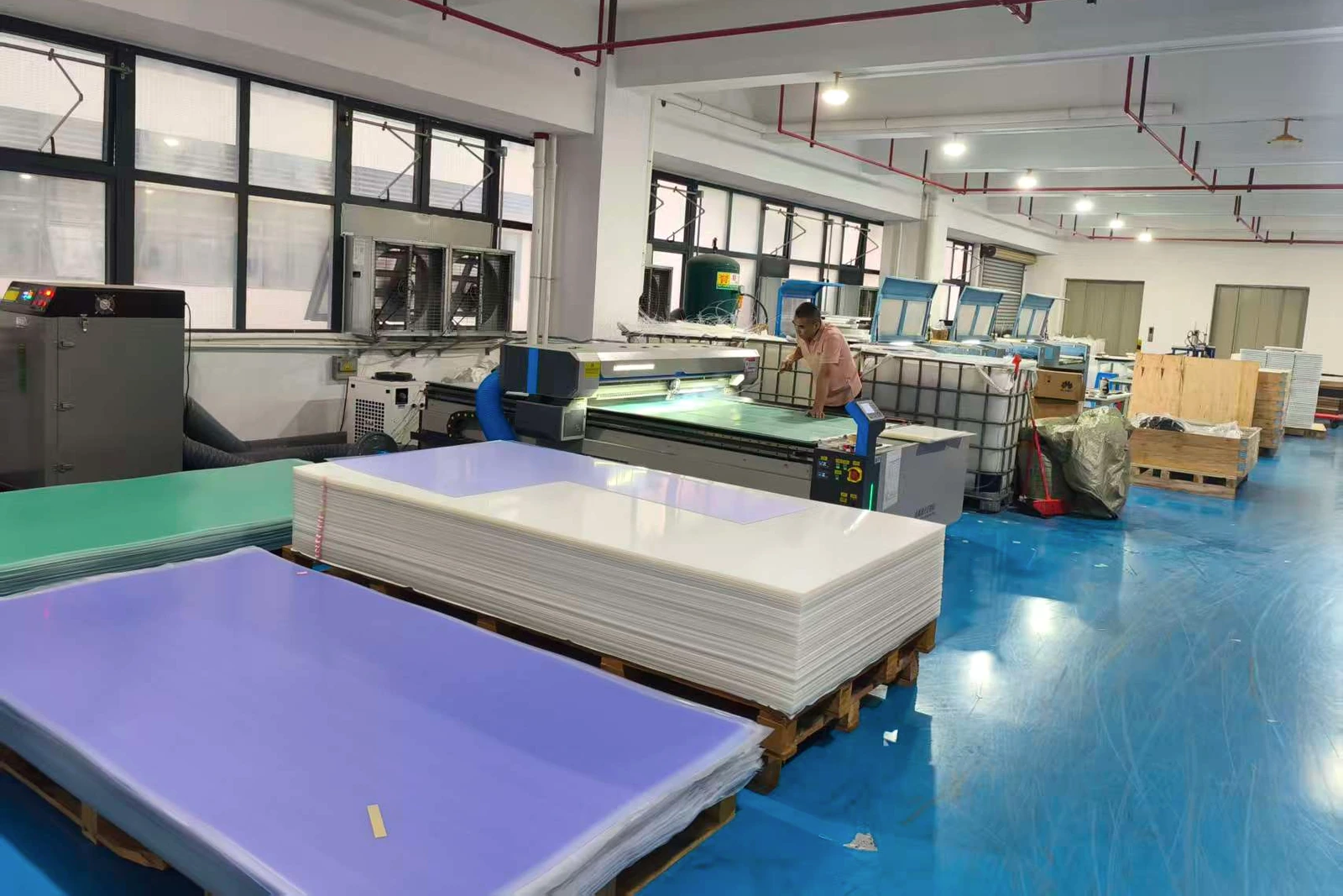 An operator stands beside processing equipment and stacks of sheet materials in a factory.